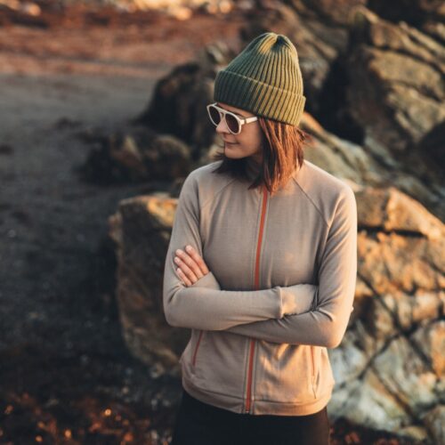 Osoba w zielonej czapce, okularach przeciwsłonecznych i beżowej kurtce zapinanej na zamek stoi na zewnątrz ze skrzyżowanymi rękami, przed dużymi skałami i rozmytym, ziemistym tłem.