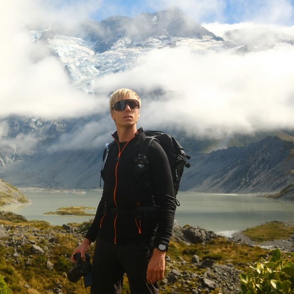 Osoba w okularach przeciwsłonecznych i sprzęcie outdoorowym stoi przed jeziorem z górami i śniegiem w tle, trzymając lornetkę. Chmury częściowo zakrywają szczyty. Scena jest jasna i malownicza.