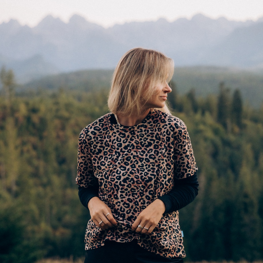 Osoba o blond włosach ubrana w koszulkę z nadrukiem lamparta stoi na zewnątrz, patrząc w bok, z lasem i górami w tle.