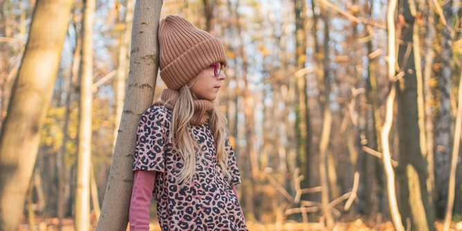Młoda dziewczyna ubrana w koszulę w panterkę, różowe długie rękawy, brązową dzianinową czapkę i okulary stoi przy drzewie w słonecznym jesiennym lesie, patrząc w bok.