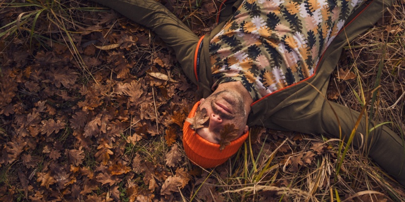 Osoba w pomarańczowej czapce i wzorzystej koszuli leży na jesiennych liściach z dwoma liśćmi zakrywającymi oczy, z wyciągniętymi rękami, otoczona suchą trawą i brązowymi liśćmi.