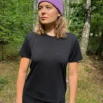 Osoba ubrana w czarną koszulkę damską z wełny merino, czarne spodnie i fioletową czapkę stoi w lesie z drzewami i zielenią w tle, patrząc lekko w bok. Światło słoneczne przesącza się przez liście, rzucając na scenę cętkowane światło.
