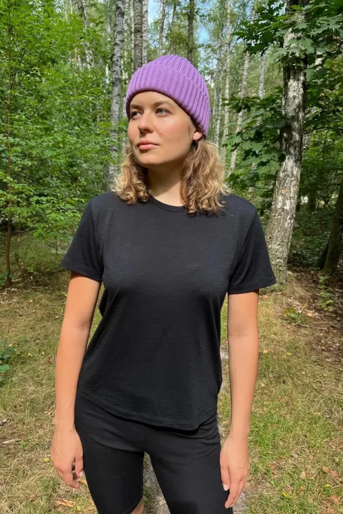 Osoba ubrana w czarną koszulkę damską z wełny merino, czarne spodnie i fioletową czapkę stoi w lesie z drzewami i zielenią w tle, patrząc lekko w bok. Światło słoneczne przesącza się przez liście, rzucając na scenę cętkowane światło.