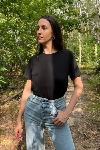 Osoba z długimi, ciemnymi włosami stoi na leśnej ścieżce, ubrana w czarny T-shirt z wełny merino i jasnoniebieskie dżinsy z wysokim stanem. Patrzy w bok, otoczona zielonymi drzewami i rozproszonym światłem słonecznym.