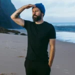 Brodaty mężczyzna w czarnym T-shircie z wełny merino męski i niebieskiej czapce stoi na plaży, osłaniając oczy i patrząc w dal. Ocean i klify stanowią tło pod zachmurzonym niebem, podkreślając męską scenę.