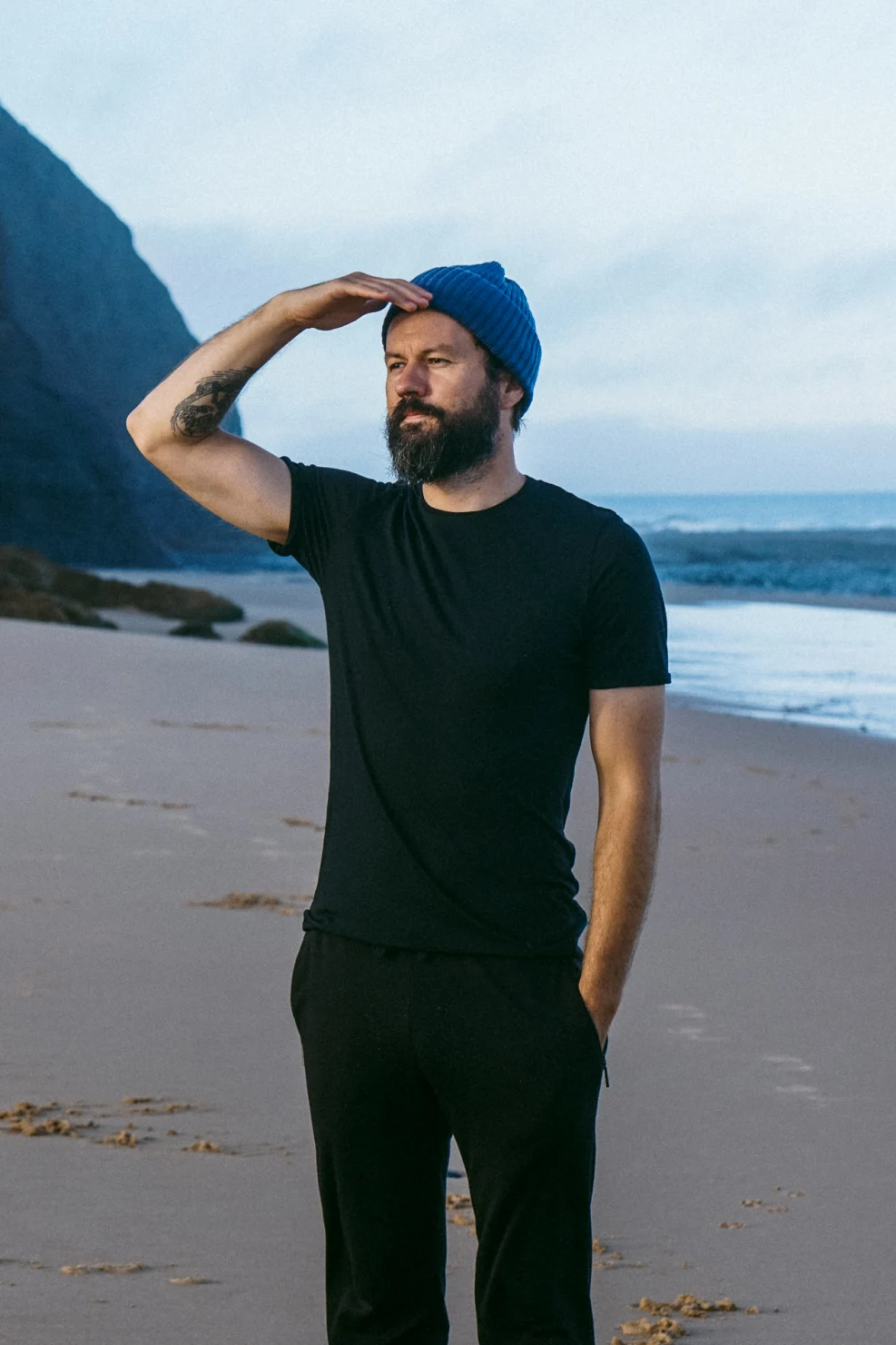 Brodaty mężczyzna w czarnym T-shircie z wełny merino męski i niebieskiej czapce stoi na plaży, osłaniając oczy i patrząc w dal. Ocean i klify stanowią tło pod zachmurzonym niebem, podkreślając męską scenę.