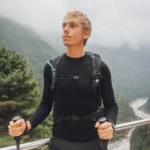 Niska, blondwłosa osoba stoi na balkonie, trzymając kije trekkingowe, ubrana w czarną bieliznę z wełny merino z długim rękawem i plecak. Za nimi mgliste góry, bujne zielone drzewa i rzeka w dolinie.