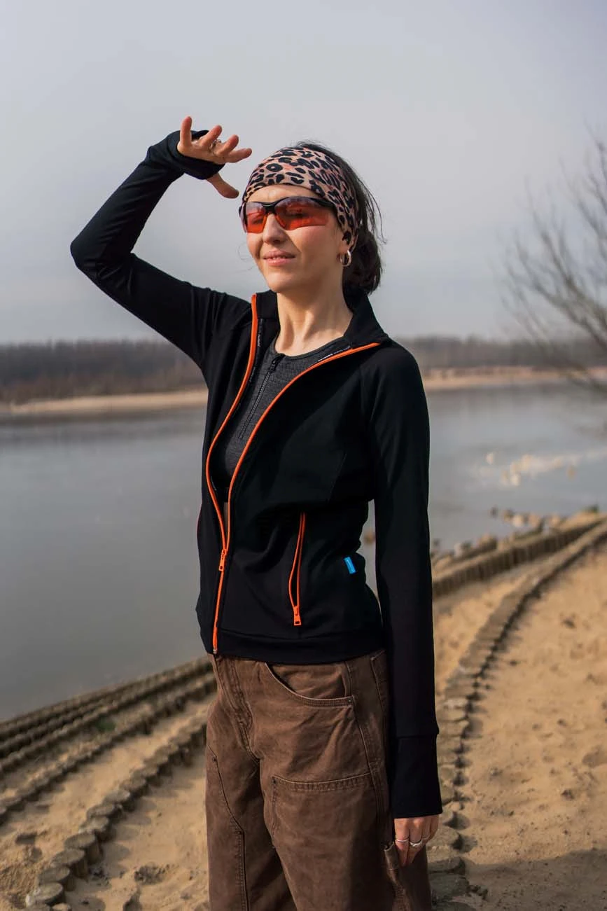 Kobieta stoi nad rzeką pod zachmurzonym niebem, ma na sobie opaskę z nadrukiem w panterkę, pomarańczowe okulary przeciwsłoneczne i czarną bluzę sportową z wełny merino z pomarańczowym wykończeniem.