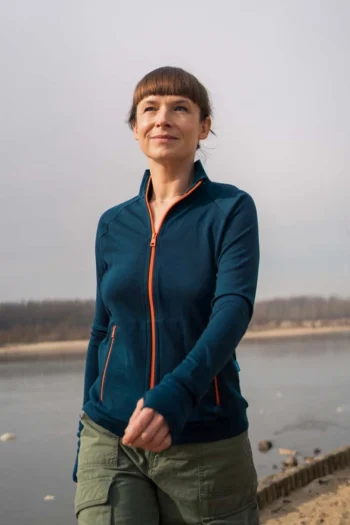 Brązowowłosa kobieta spaceruje na zewnątrz wzdłuż rzeki, ubrana w sportową, rozpinaną bluzę z wełny merynosów w kolorze petrol blue z pomarańczowymi wykończeniami i oliwkowozielone spodnie pod zachmurzonym niebem.