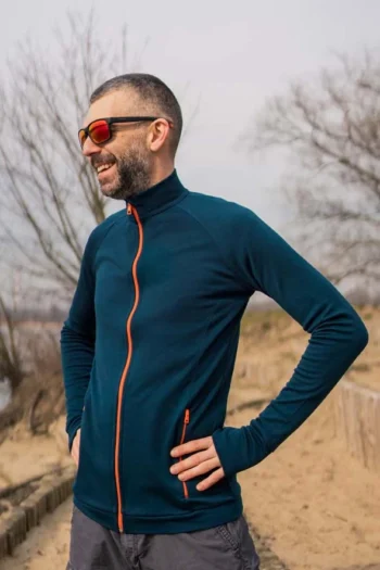 Na piaszczystej ścieżce stoi pewnie osoba ubrana w bluzę z wełny merino sportową męską petrol z pomarańczowymi akcentami, szare szorty i okulary przeciwsłoneczne w odcieniu czerwieni.