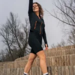 Kobieta w czarnym stroju sportowym i pomarańczowych trampkach pewnie idzie, uśmiechając się i podnosząc rękę. Stoi na drewnianych schodach, a za nią widać nagie drzewa, co wskazuje na chłodny dzień idealny na nakładanie warstw. Jej wybór Bluzy z wełny merino sportowa damska czarna oraz kolarki z wełny merino czarna zapewnia idealne połączenie ciepła i stylu.