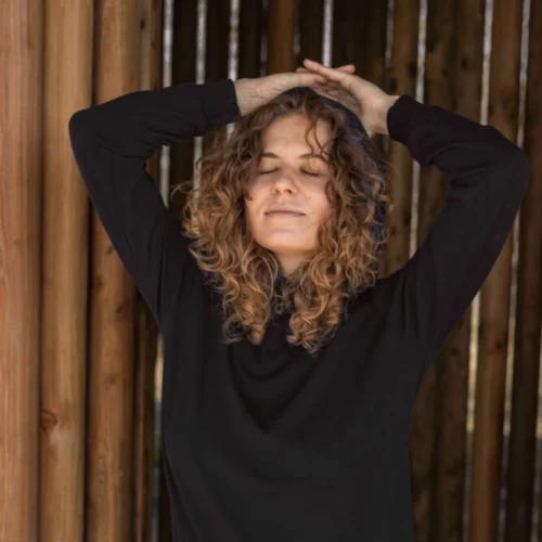 Osoba z kręconymi włosami stoi z zamkniętymi oczami i podniesionymi rękami, ubrana w Bluza z wełny merino damska z kapturem czarna i jasne spodnie przed drewnianym tłem.