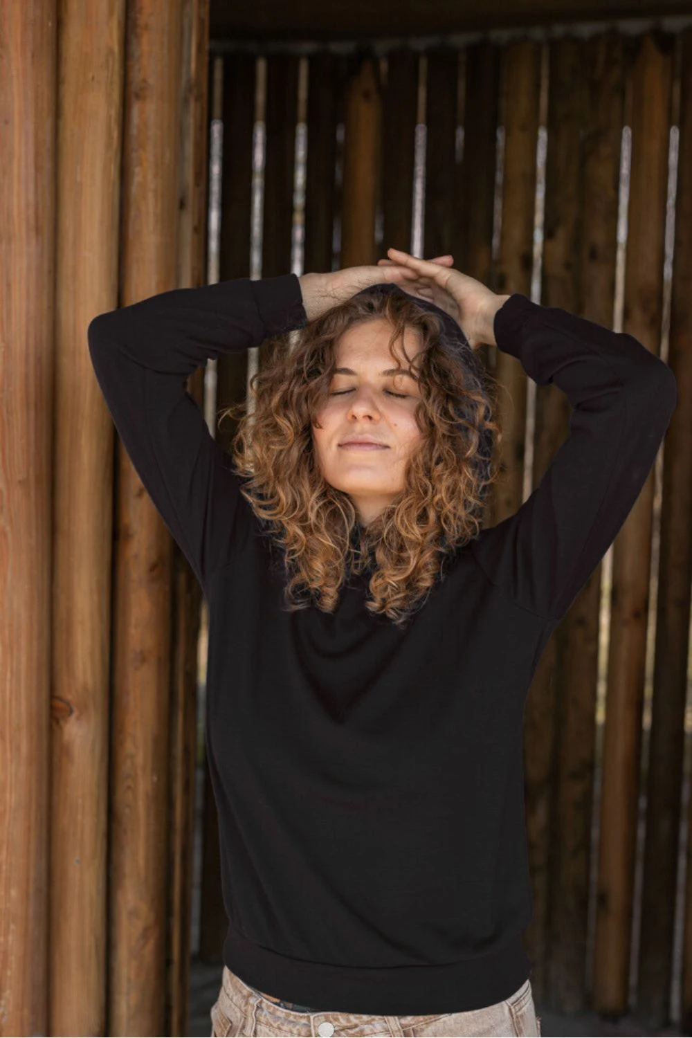Osoba z kręconymi włosami stoi z zamkniętymi oczami i podniesionymi rękami, ubrana w Bluza z wełny merino damska z kapturem czarna i jasne spodnie przed drewnianym tłem.
