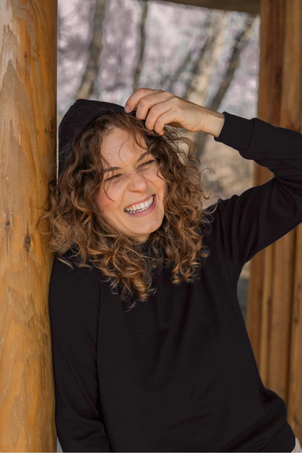 Młoda kobieta z kręconymi włosami uśmiecha się, naciągając kaptur na głowę, ubrana w Bluzę z wełny merino damską z kapturem czarną. Opiera się o drewniany słup z rozmytymi drzewami i miękkim światłem zewnętrznym w tle.