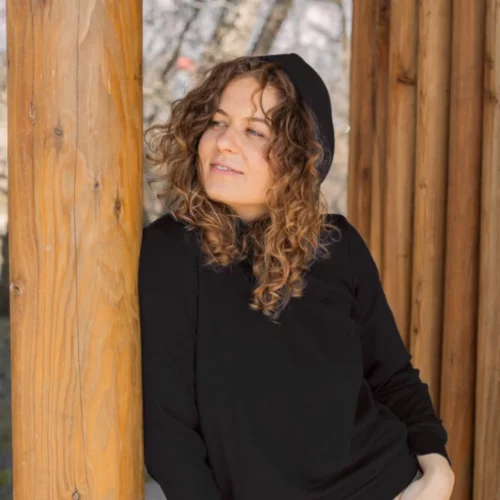 Kobieta z kręconymi włosami relaksuje się na zewnątrz w bluzie z kapturem "Bluza z wełny merino damska czarna" i beżowych spodniach, opierając się o drewniany słup i patrząc w bok.