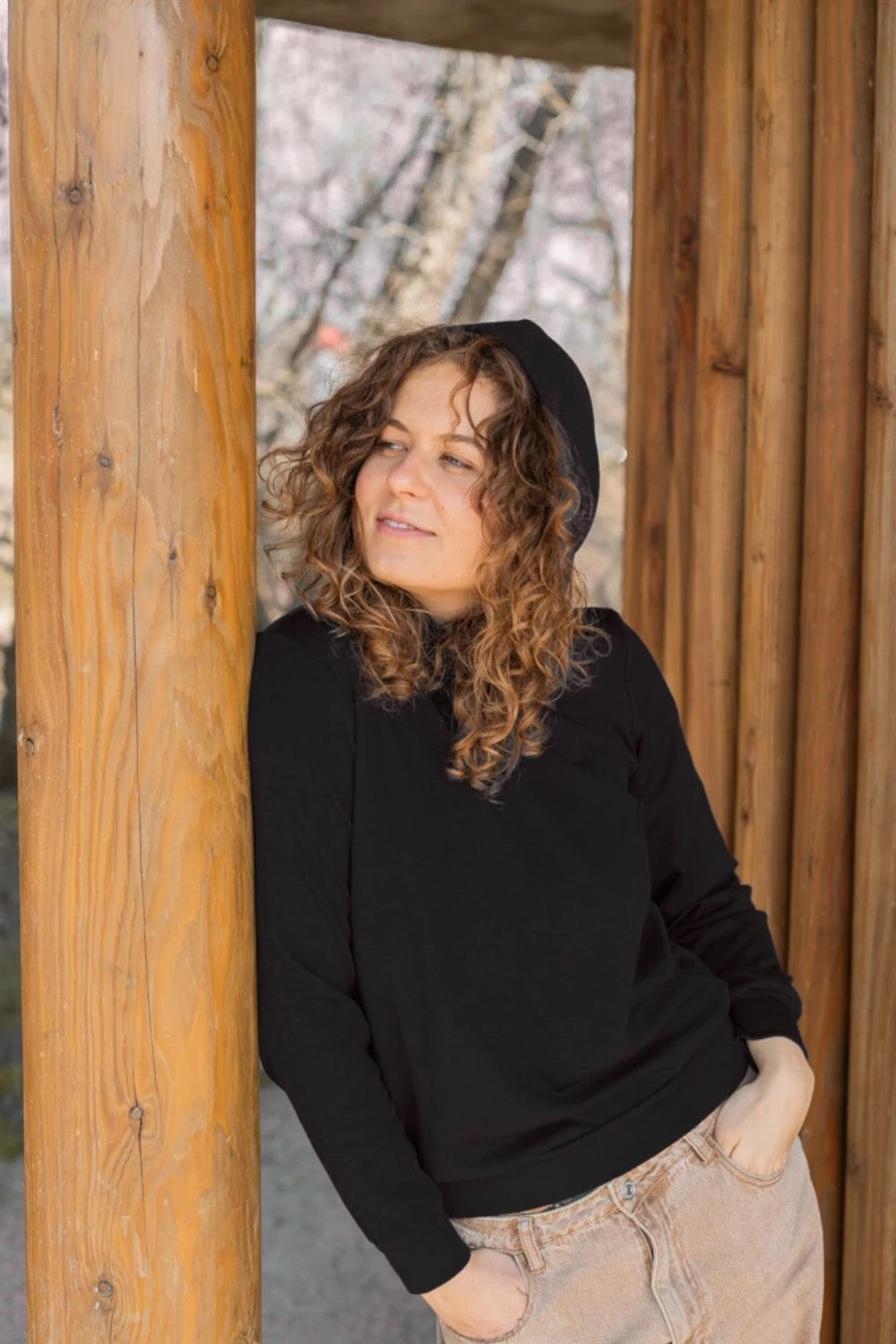 Kobieta z kręconymi włosami relaksuje się na zewnątrz w bluzie z kapturem "Bluza z wełny merino damska czarna" i beżowych spodniach, opierając się o drewniany słup i patrząc w bok.