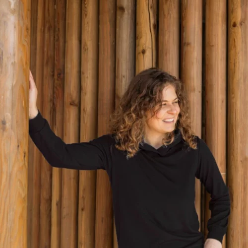 Kobieta z kręconymi włosami ubrana w Bluzę z wełny merino damską z kapturem czarną i beżowe spodnie opiera się o drewniane słupy, uśmiechając się i patrząc w bok.