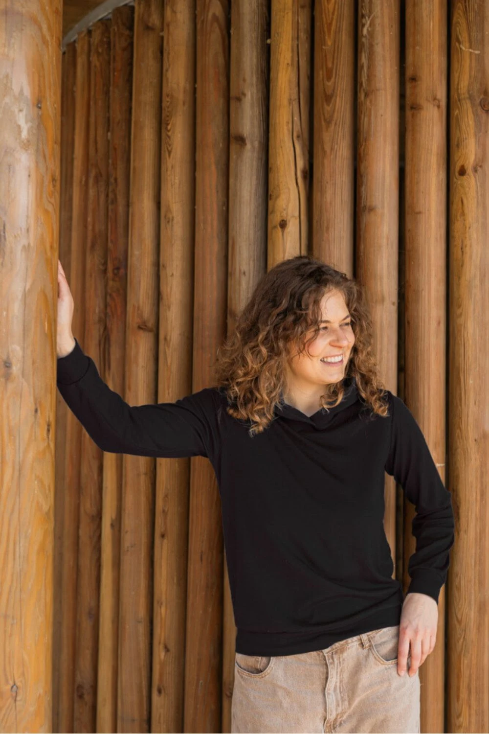 Kobieta z kręconymi włosami ubrana w Bluzę z wełny merino damską z kapturem czarną i beżowe spodnie opiera się o drewniane słupy, uśmiechając się i patrząc w bok.