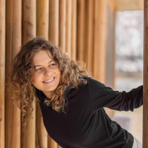 Młoda kobieta z kręconymi włosami, ubrana w czarną bluzę z kapturem Bluza z wełny merino damska, pochyla się figlarnie między drewnianymi filarami i uśmiecha w bok, z delikatnie rozmytym tłem na zewnątrz.