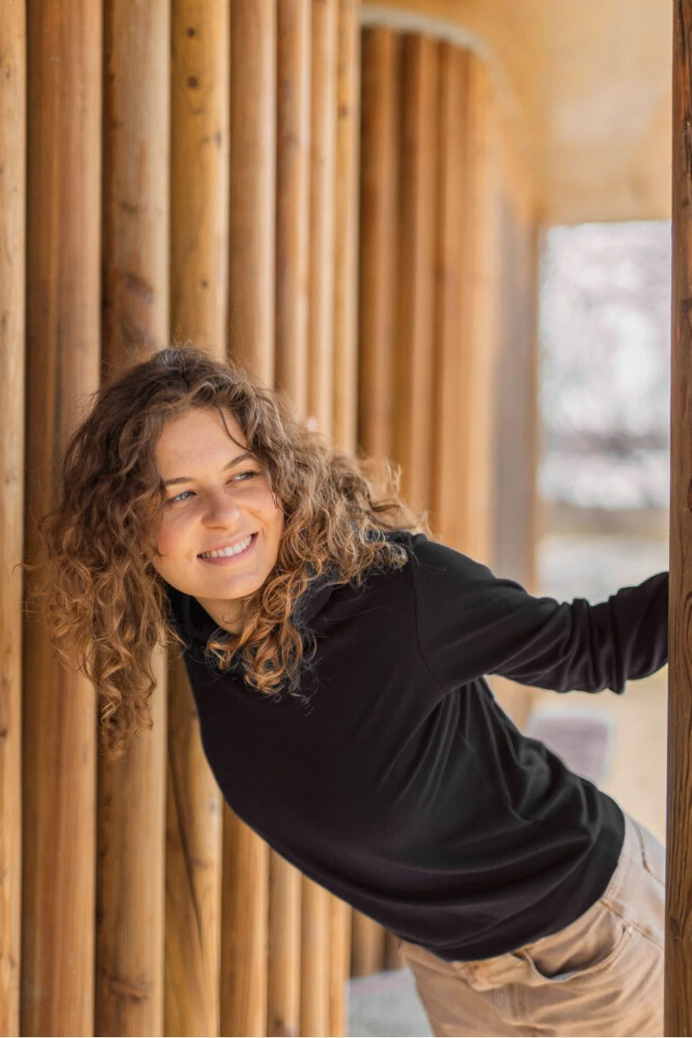 Młoda kobieta z kręconymi włosami, ubrana w czarną bluzę z kapturem Bluza z wełny merino damska, pochyla się figlarnie między drewnianymi filarami i uśmiecha w bok, z delikatnie rozmytym tłem na zewnątrz.