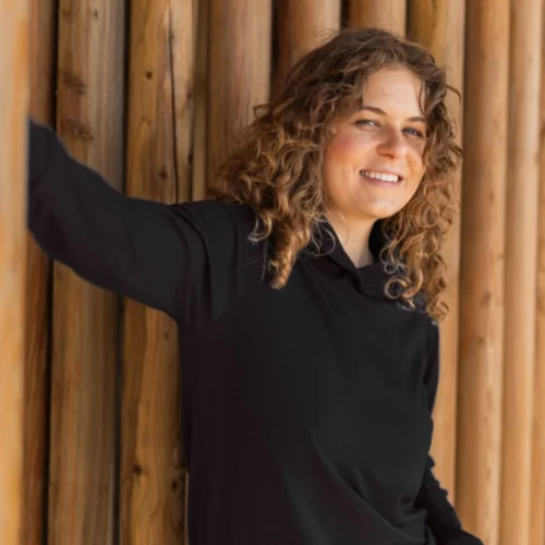 Kobieta z kręconymi włosami, uśmiechnięta do kamery, opiera się o ścianę z pionowych drewnianych bali, ubrana w Bluza z wełny merino damska z kapturem czarna i lekkie spodnie.