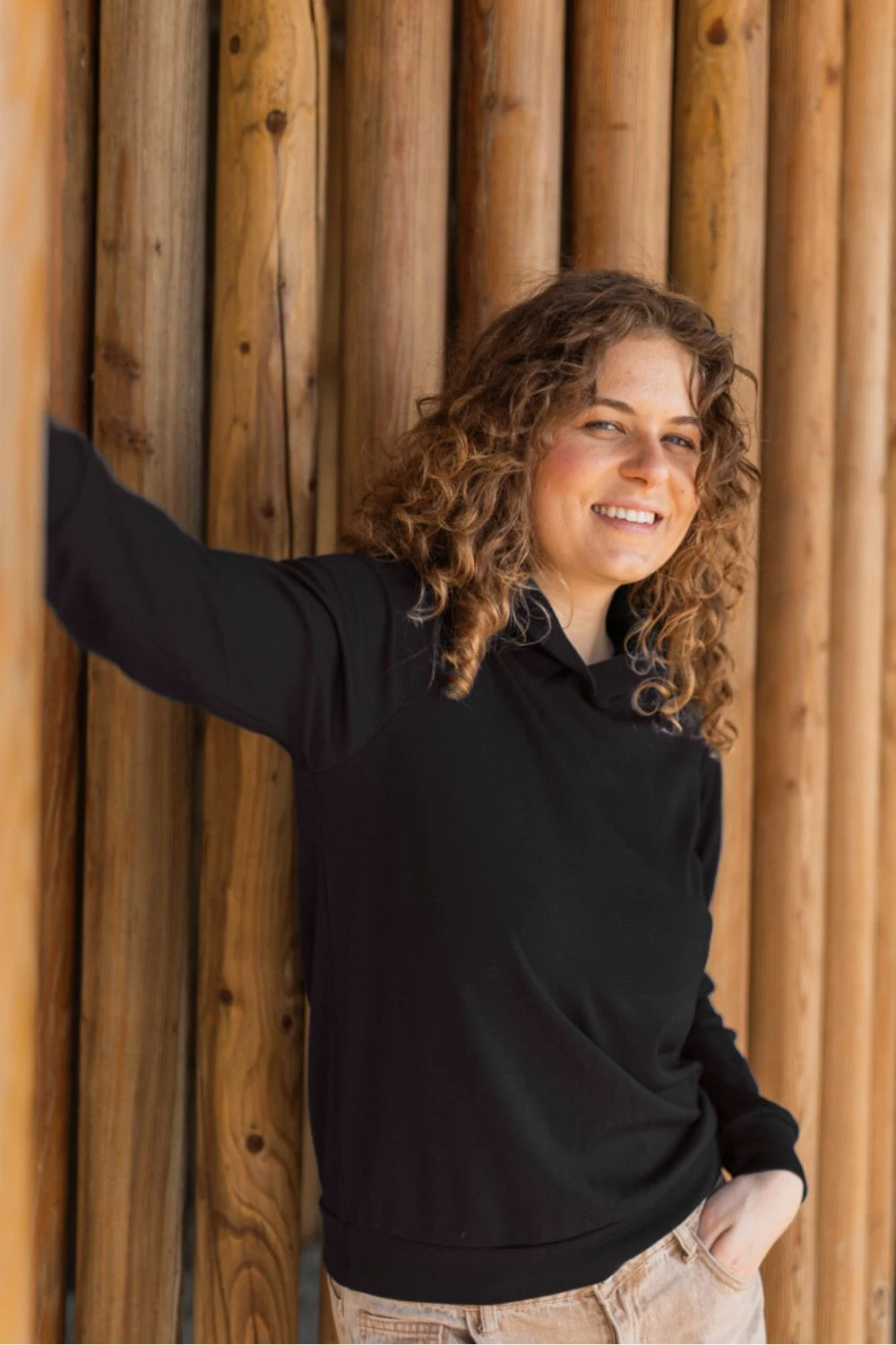 Kobieta z kręconymi włosami, uśmiechnięta do kamery, opiera się o ścianę z pionowych drewnianych bali, ubrana w Bluza z wełny merino damska z kapturem czarna i lekkie spodnie.