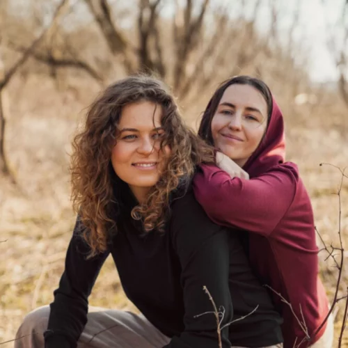 Dwie kobiety siedzą na zewnątrz w bezlistnym lesie. Kobieta z przodu, uśmiechnięta, ma kręcone włosy i nosi "Bluzę z wełny merino damską z kapturem czarną". Za nią inna kobieta w bordowej bluzie z kapturem opiera się o jej plecy, obie uśmiechają się ciepło.