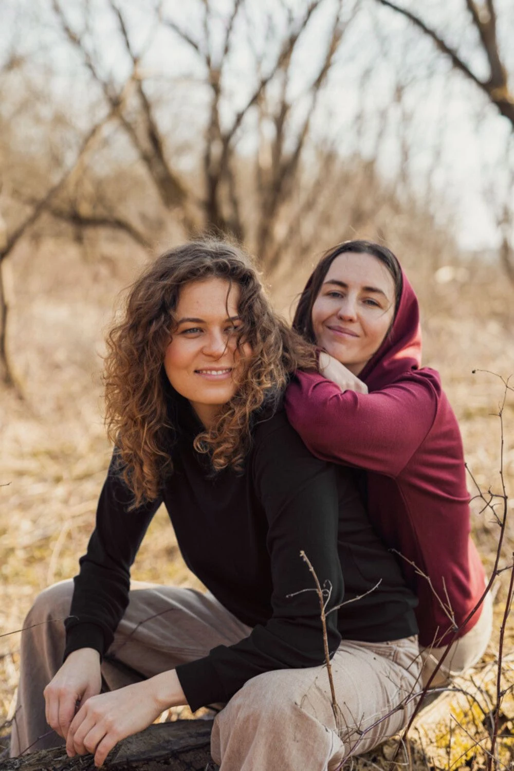 Dwie kobiety siedzą na zewnątrz w bezlistnym lesie. Kobieta z przodu, uśmiechnięta, ma kręcone włosy i nosi "Bluzę z wełny merino damską z kapturem czarną". Za nią inna kobieta w bordowej bluzie z kapturem opiera się o jej plecy, obie uśmiechają się ciepło.