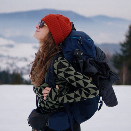 Kobieta ubrana w ceglastoczerwoną czapkę z wełny merynosów, okulary przeciwsłoneczne i wzorzysty sweter stoi z dużym niebieskim plecakiem na tle zaśnieżonego krajobrazu z górami i drzewami w tle.