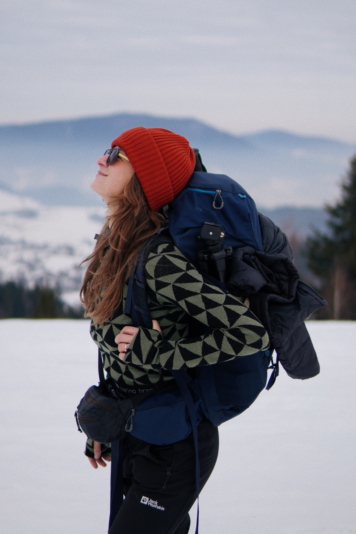 Kobieta ubrana w ceglastoczerwoną czapkę z wełny merynosów, okulary przeciwsłoneczne i wzorzysty sweter stoi z dużym niebieskim plecakiem na tle zaśnieżonego krajobrazu z górami i drzewami w tle.