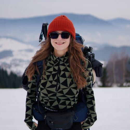 Uśmiechnięta osoba w Czapce z wełny merino ściągaczowej, okularach przeciwsłonecznych i wzorzystej kurtce stoi w śnieżnym krajobrazie z górami i drzewami, niosąc duży plecak turystyczny.