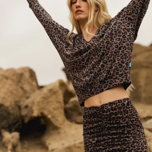 Kobieta z długimi blond włosami stoi na zewnątrz na skalistym terenie, z podniesionymi rękami i zamkniętymi oczami, ubrana w Spódnicę z wełny merino marszczona szara panterka - marszczoną spódnicę z wełny merino w szary lamparci print.