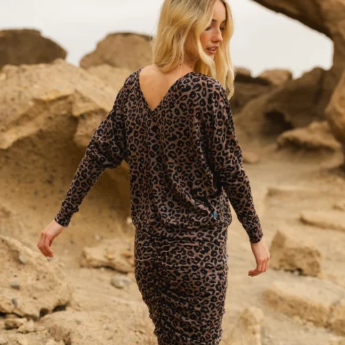 Kobieta o długich blond włosach modeluje "Spódnicę z wełny merynos marszczona szara panterka", spacerując na świeżym powietrzu po skalistym terenie z dużymi skałami i zachmurzonym niebem w tle, spoglądając przez ramię.