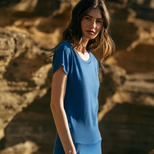 Kobieta z długimi brązowymi włosami stoi na zewnątrz przed skalistym, oświetlonym słońcem terenem, ubrana w Spódnicę z wełny merino marszczona szara panterka i patrzy w kamerę.