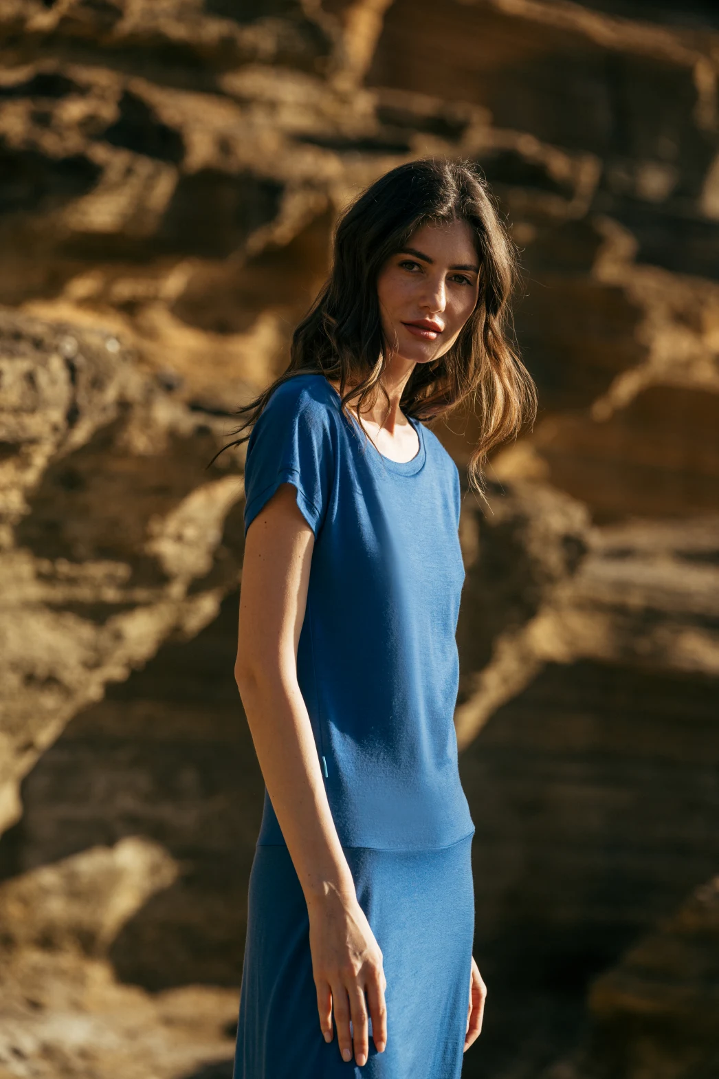 Kobieta z długimi brązowymi włosami stoi na zewnątrz przed skalistym, oświetlonym słońcem terenem, ubrana w Spódnicę z wełny merino marszczona szara panterka i patrzy w kamerę.