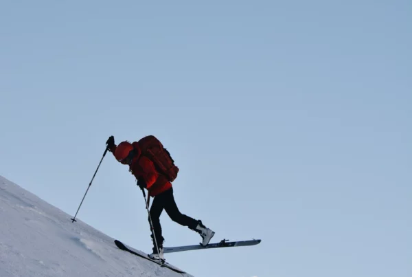 Osoba ubrana w czerwony sprzęt zimowy i plecak używa kijków narciarskich, aby wspiąć się na strome, ośnieżone zbocze z ośnieżonymi górami w tle pod czystym niebem.