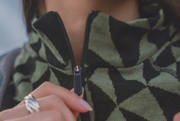 Zbliżenie na osobę zapinającą zielono-czarną kurtkę z geometrycznym wzorem, z naciskiem na zamek błyskawiczny i kołnierz; widoczna jest część dłoni i srebrny pierścionek.