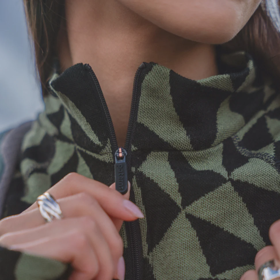 Zbliżenie na osobę zapinającą zielono-czarną kurtkę z geometrycznym wzorem, z naciskiem na zamek błyskawiczny i kołnierz; widoczna jest część dłoni i srebrny pierścionek.