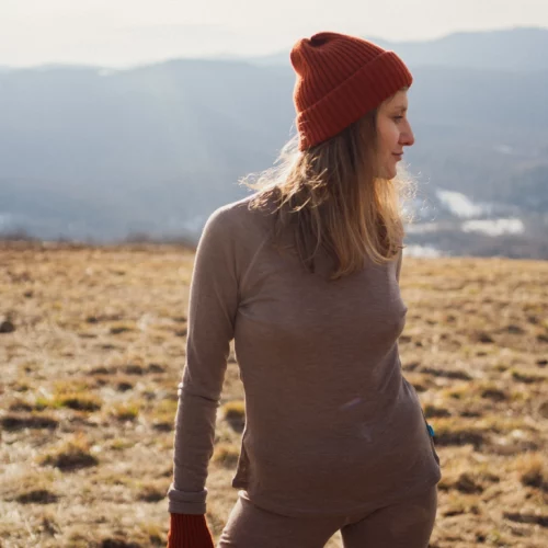 Osoba ubrana w Dwupak: Bielizna damska góra i legginsy beżowy melanż stoi na trawiastym polu z górami i zamglonym niebem za nimi, patrząc w prawo, gdy światło słoneczne oświetla scenę.