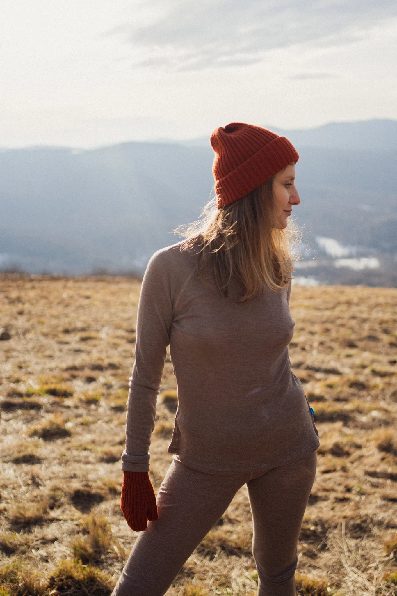 Osoba ubrana w Dwupak: Bielizna damska góra i legginsy beżowy melanż stoi na trawiastym polu z górami i zamglonym niebem za nimi, patrząc w prawo, gdy światło słoneczne oświetla scenę.