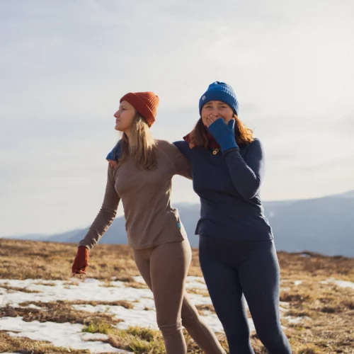 Dwie kobiety noszą Dwupak: Bielizna damska góra i legginsy beżowy melanż, uśmiechając się, gdy spacerują na świeżym powietrzu po trawiastym, zaśnieżonym zboczu i podziwiają malowniczy górski krajobraz w tle.