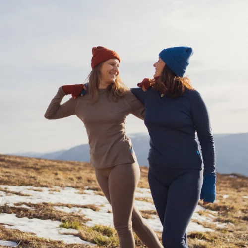 Dwie kobiety ubrane w Dwupak: Bielizna damska góra i legginsy beżowy melanż uśmiechają się i figlarnie pozują na świeżym powietrzu na ośnieżonym zboczu z górami w tle pod czystym niebem.