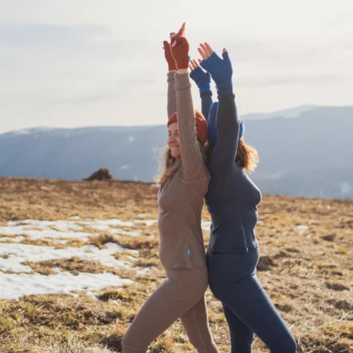 Dwie osoby ubrane w Dwupak: Bielizna damska góra i legginsy beżowy melanż stoją tyłem do siebie na trawiastym, zaśnieżonym polu, unosząc radośnie ramiona z górami i jasnym niebem w tle.