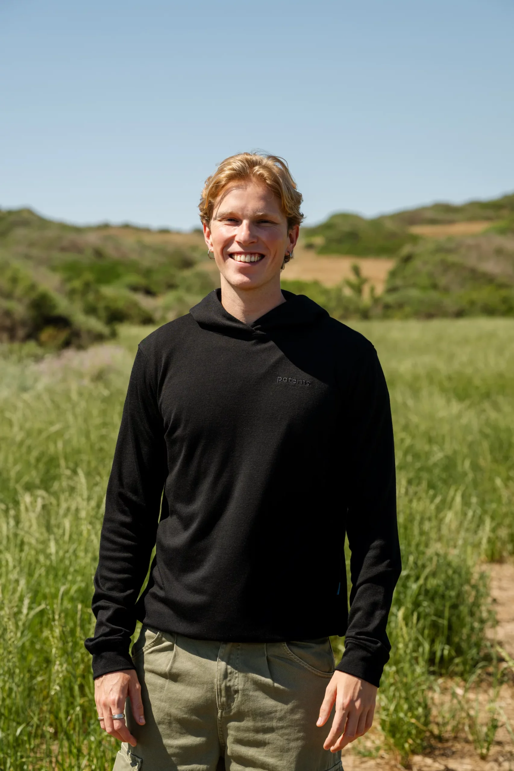Uśmiechnięty młody mężczyzna o blond włosach stoi na zewnątrz na trawiastym polu ubrany w oliwkową męską bluzę przeciwsłoneczną z wełny merynosów, z zielonymi wzgórzami i czystym błękitnym niebem w tle.