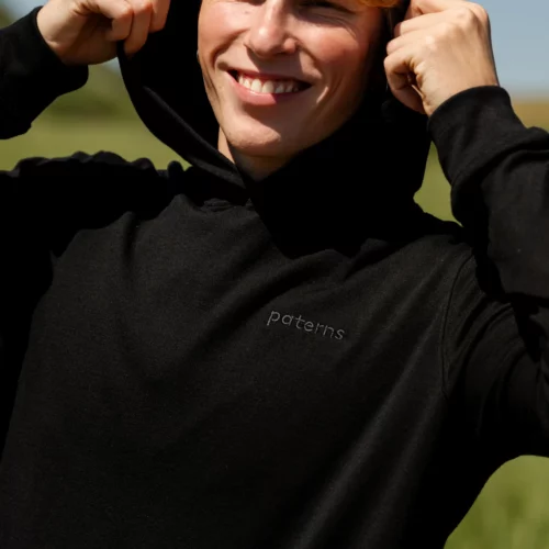Uśmiechnięta osoba o blond włosach zakłada męską oliwkową bluzę Bluza sunhoodie z wełny merino. Bluza posiada napis "paterns" na klatce piersiowej. Są na zewnątrz z rozmytą zielenią i błękitnym niebem w tle.