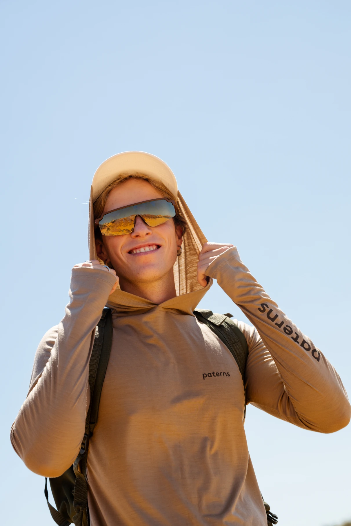 Osoba ubrana w Bluza przeciwsłoneczna z wełny merino męska oliwkowa, okulary przeciwsłoneczne i beżowy kapelusz uśmiecha się na zewnątrz pod czystym, błękitnym niebem.