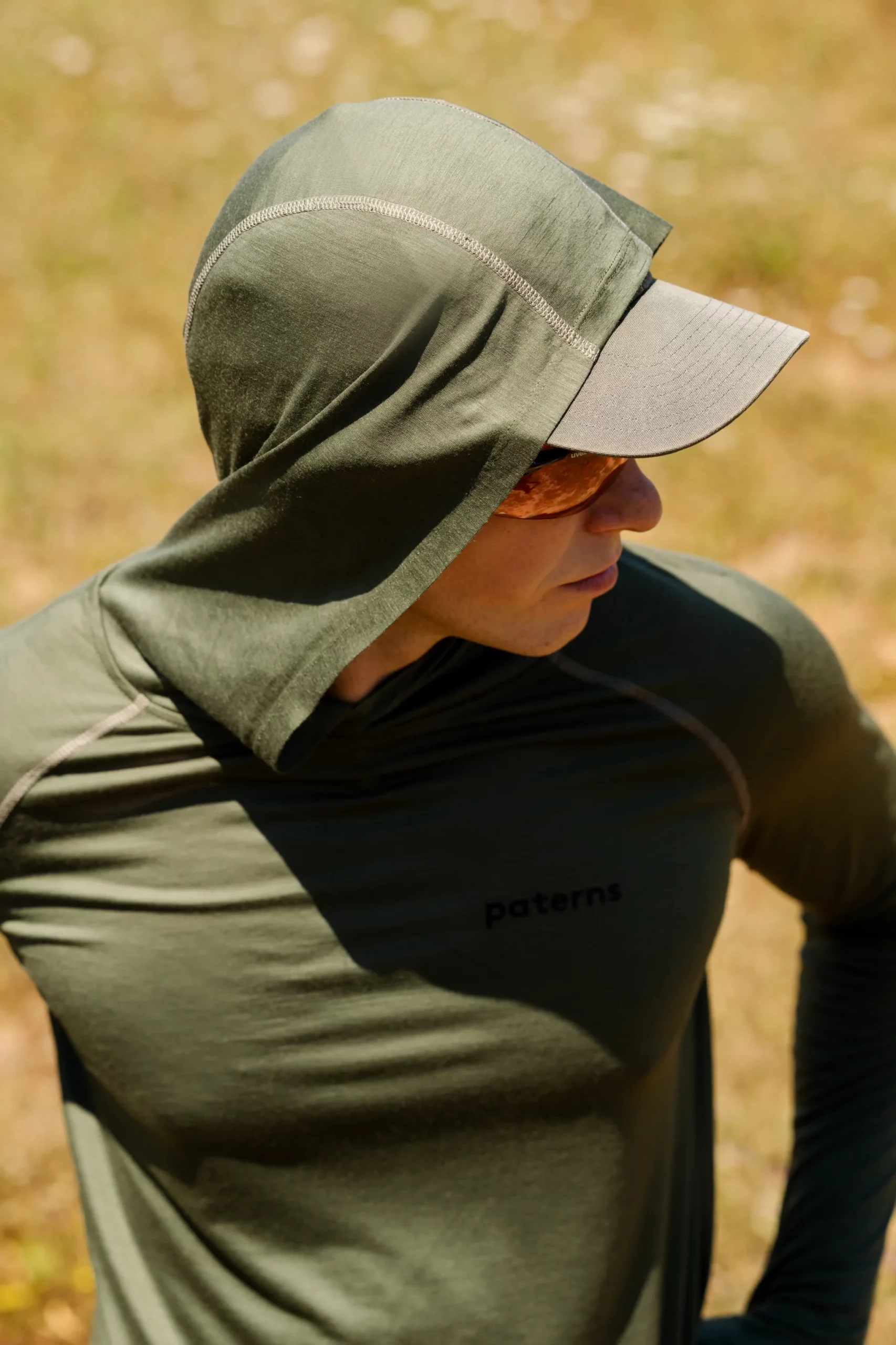 Mężczyzna w zielonej bluzie przeciwsłonecznej "Bluza z wełny merino męska oliwkowa", okularach przeciwsłonecznych i szarej czapce stoi na zewnątrz na trawie, patrząc w bok. Bluza z kapturem ma nadruk "paterns" na piersi.
