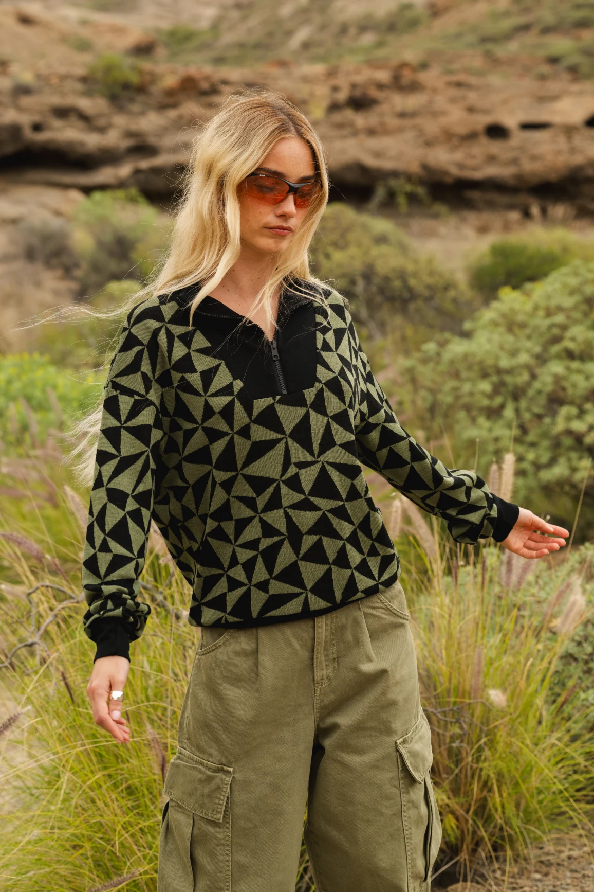 Kobieta o długich blond włosach, ubrana w okulary przeciwsłoneczne, czarno-zielony sweter w geometryczne wzory i oliwkowe spodnie cargo, stoi na zewnątrz w trawiastym, skalistym krajobrazie.