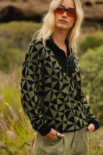 Kobieta z długimi blond włosami nosi pomarańczowe okulary przeciwsłoneczne, czarno-zielony sweter w geometryczne wzory i spodnie khaki, stojąc na zewnątrz w trawiastym otoczeniu przyrody.