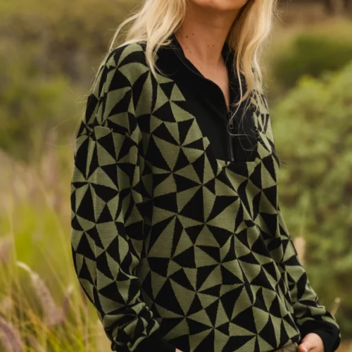 Kobieta z długimi blond włosami nosi pomarańczowe okulary przeciwsłoneczne, czarno-zielony sweter w geometryczne wzory i spodnie khaki, stojąc na zewnątrz w trawiastym otoczeniu przyrody.